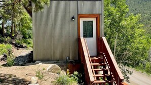 Uma cabana pequena com deck de madeira e fachada metálica, rodeada por vegetação natural e montanhas ao fundo.
