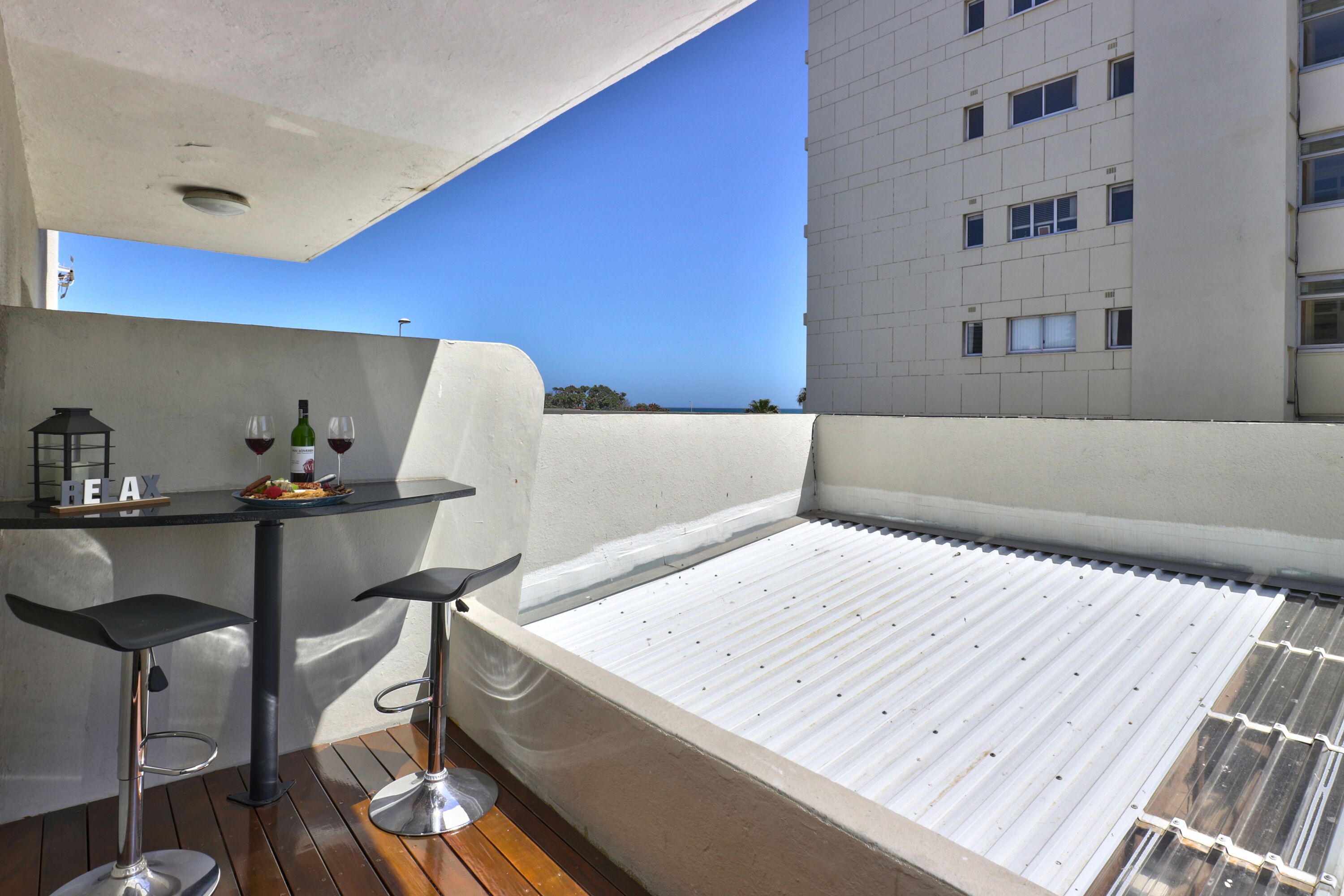 A rooftop terrace with a pool, two bar stools, a table with a bottle and glasses, and a view of a building and clear sky.
