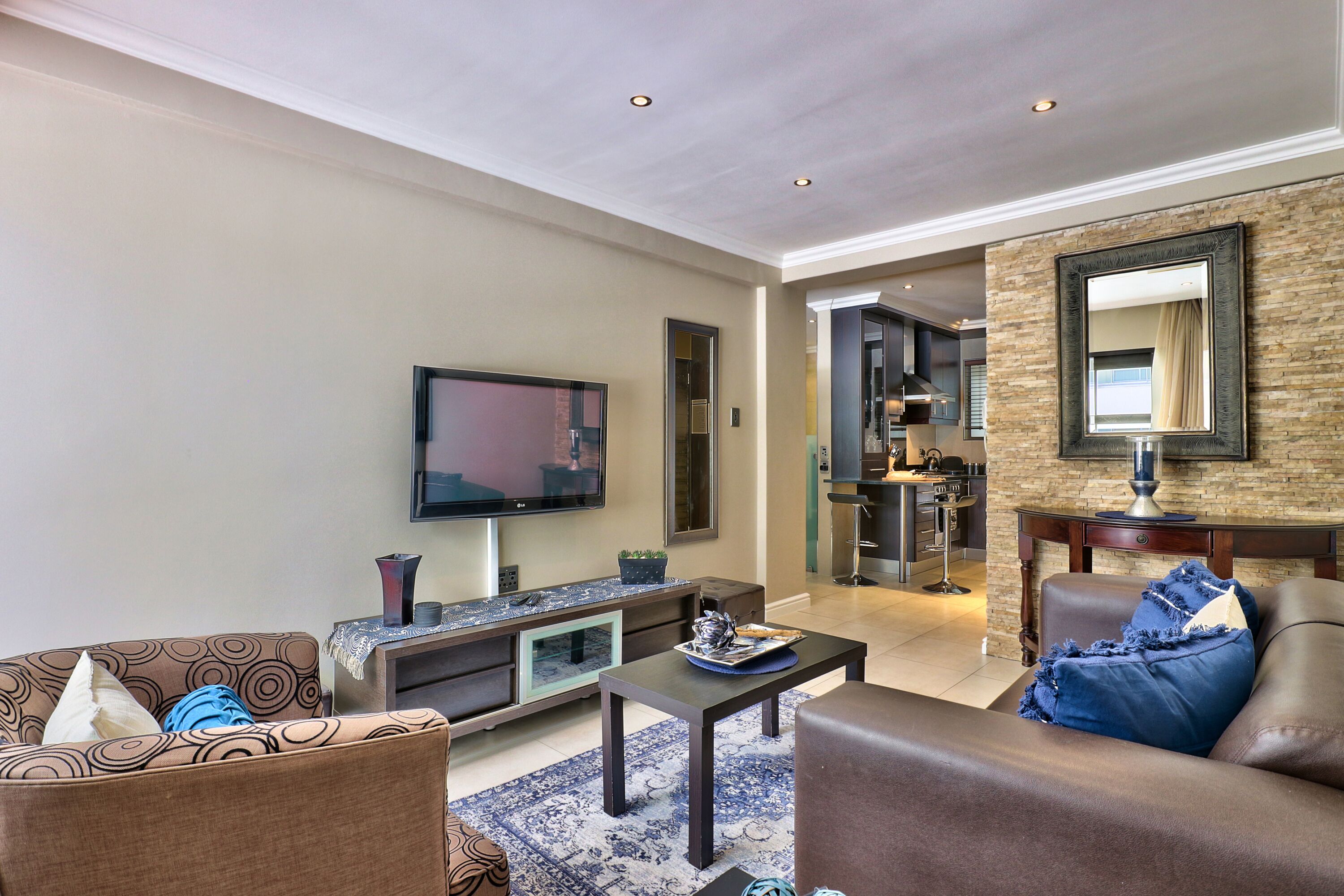A modern living room with a flat-screen TV, a sofa, a coffee table, and a kitchen area visible in the background.