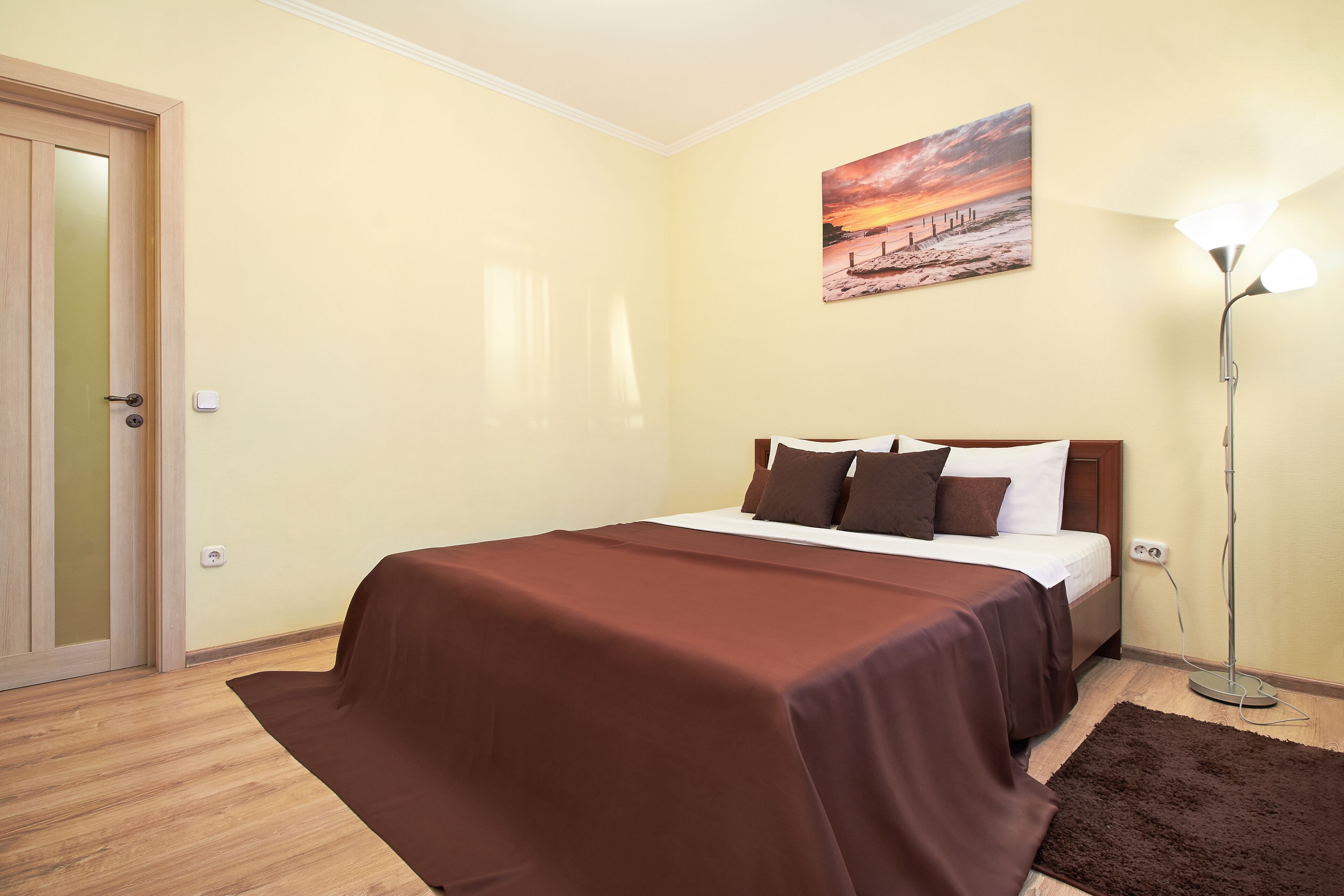 A bedroom with a bed, a painting on the wall, a floor lamp, and a door.
