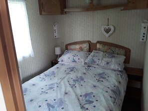 A bedroom with a bed, wooden headboard, patterned bedding, a bedside lamp, and a wall-mounted shelf. - 3 bed Caravan Suzie 1,10 Mins From Beach Suzie 1 (Hull)