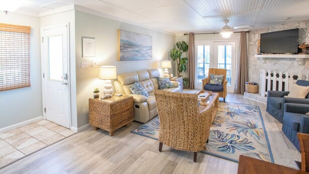 A spacious living room with a sofa, two wicker chairs, a coffee table, and a TV mounted on a stone wall. - Marooned Mariner, Tybee Island (TYBEE ISLAND)