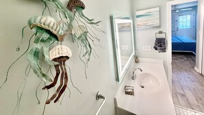 A bathroom with a white sink, a mirror, and a wall-mounted decorative piece. - Makin' Memories South Beach~OceanView 3 queen beds (Tybee Island)