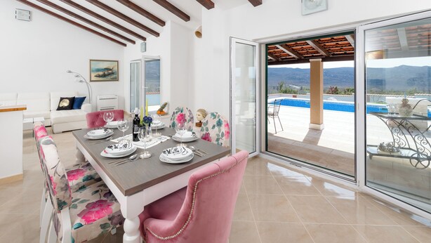 A dining area with a table set for four, a white sofa, and a view of a swimming pool and mountains through large glass doors.