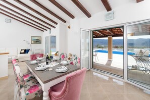 A dining area with a table set for four, a sofa, and a view of a swimming pool and mountains. - Villa Dea (Vučevica)