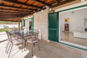An outdoor dining area with a table and chairs, a stone wall, and a view into a living room with a sofa and a cabinet. - Villa Dea (Vučevica)