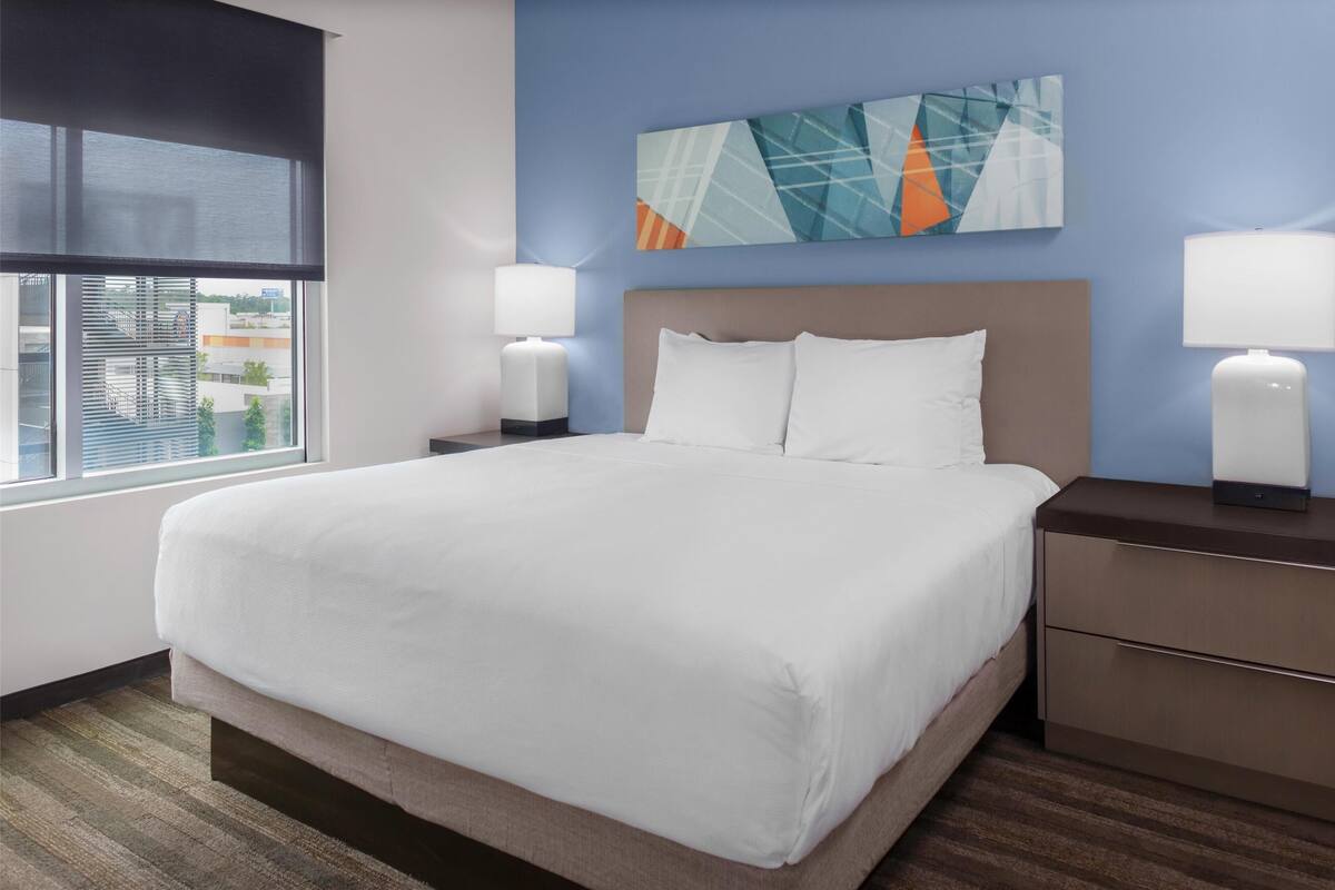 Habitación de hotel con una cama grande, dos mesitas de noche con lámparas, una ventana con persianas y vistas a la ciudad.