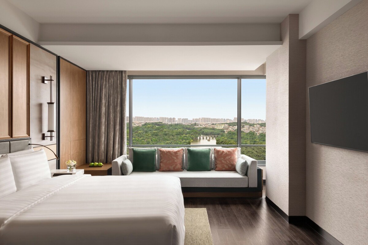 A modern hotel room with a large bed, a seating area with cushions, a TV, and a city view.