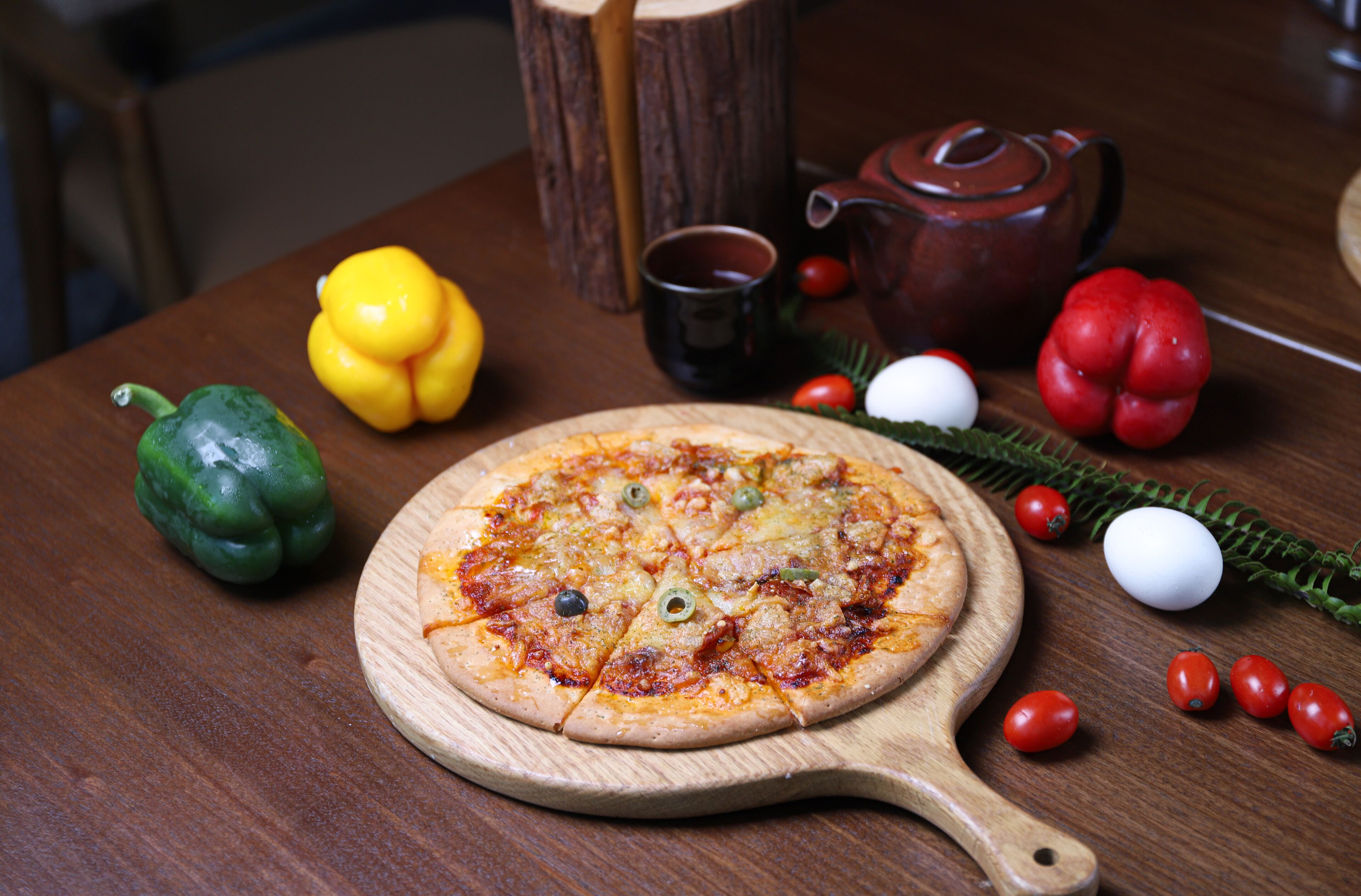 Une pizza servie sur une planche en bois dotée d’une anse en bois, entourée de divers légumes et accompagnée d’une théière posée sur une table en bois.