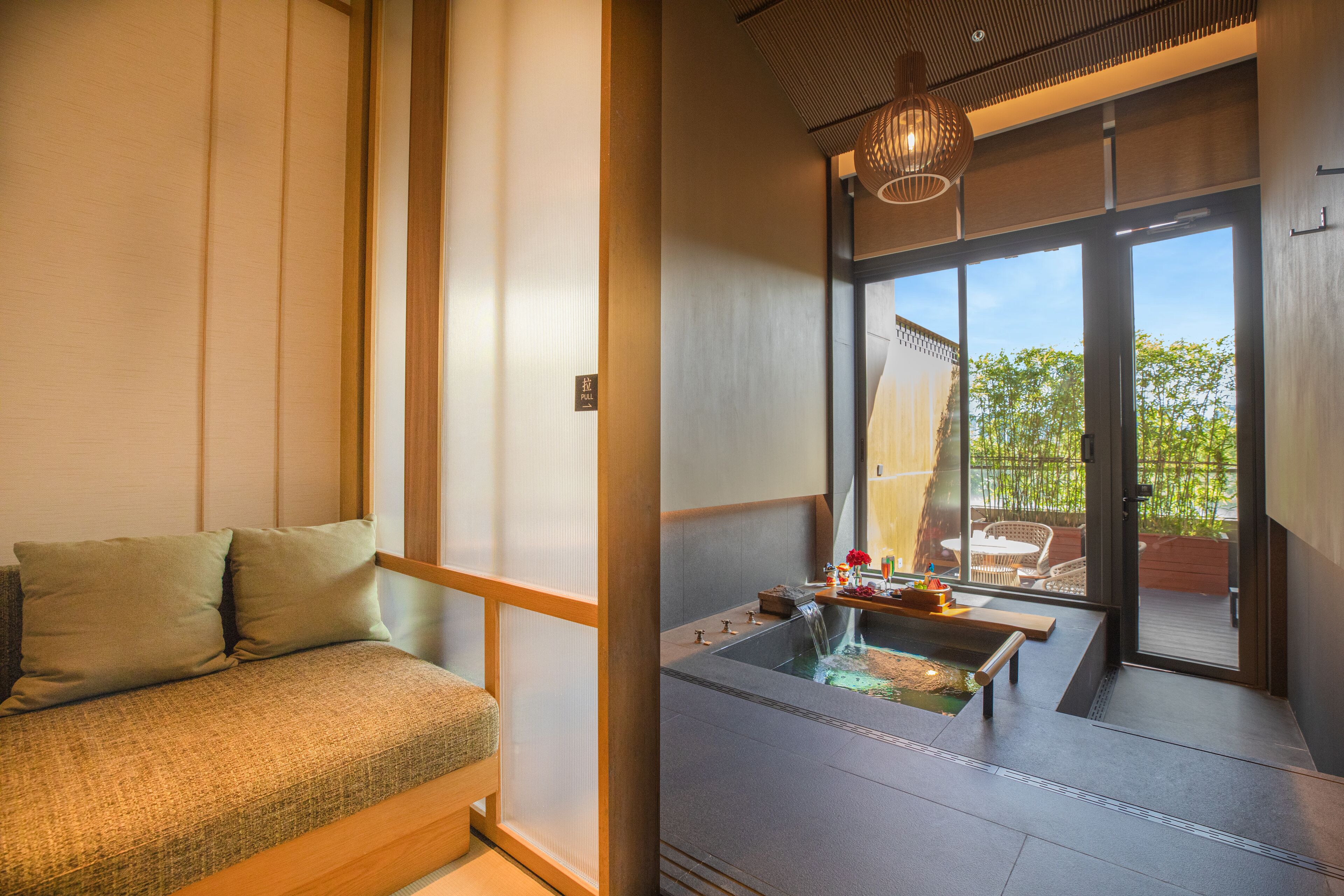 Una habitación japonesa tradicional con suelo de tatami, una mesa baja de madera y un puertas correderas que dan a un área exterior con vistas a zonas verdes.