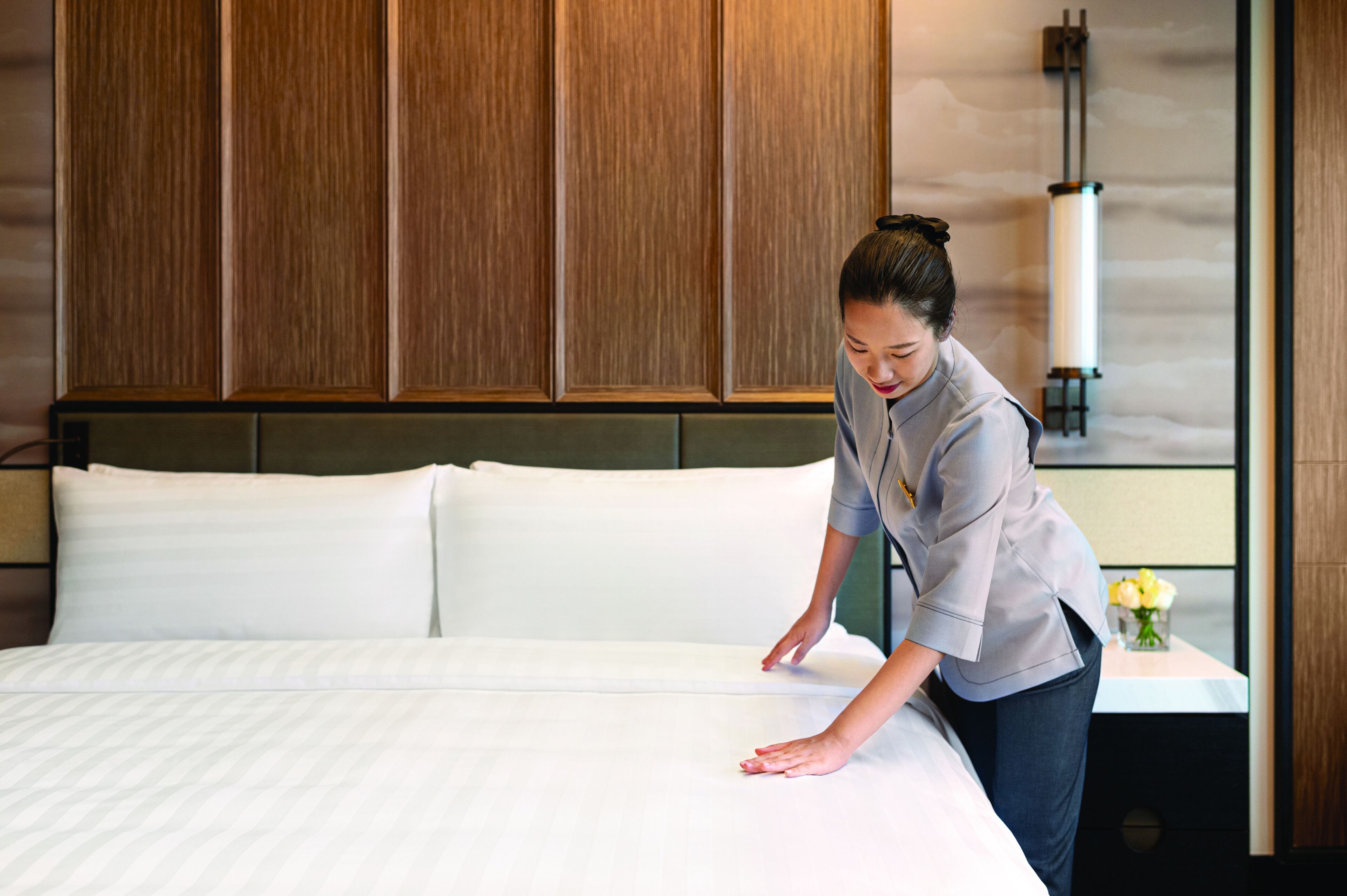 A hotel room with a neatly made bed, wooden paneling on the walls, and a bedside table with a vase of flowers.