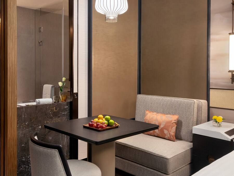A modern hotel room with a marble bathroom, a small dining table with fruit, a sofa, and a nightstand with a lamp.