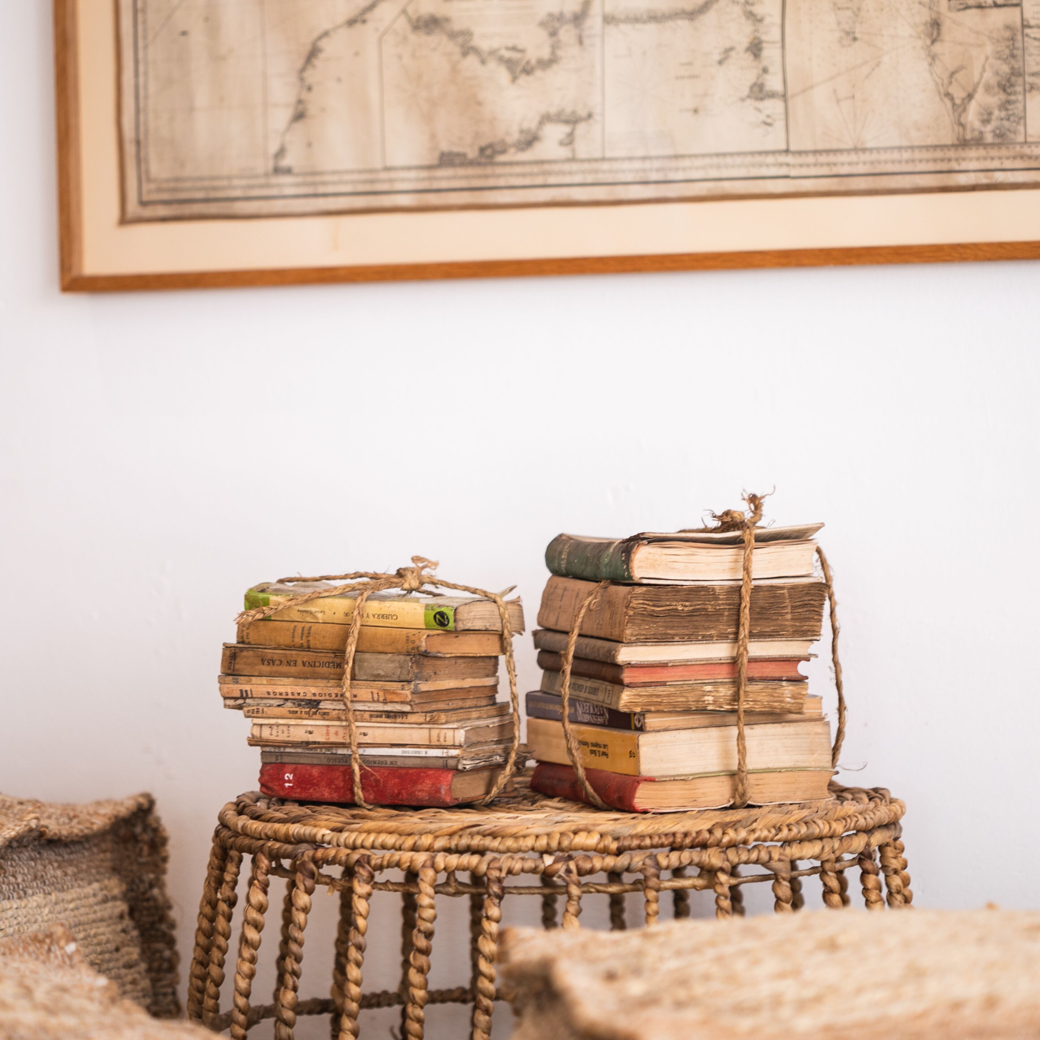 Dos cestas de mimbre apiladas con libros antiguos atados con hilo, sobre un taburete tejido frente a una pared lisa con un mapa enmarcado arriba.