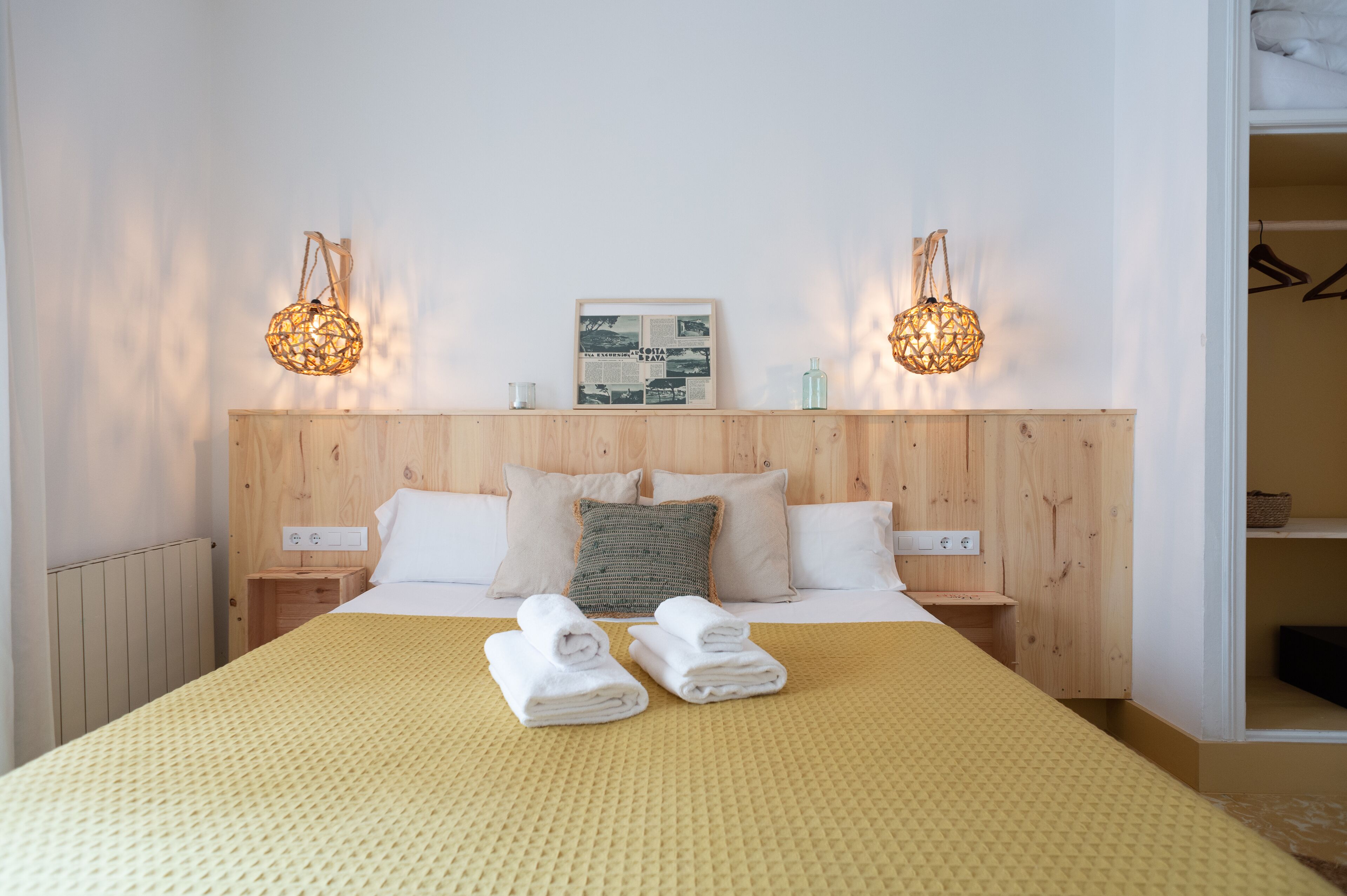 Una cama bien hecha con dos toallas enrolladas, cabecera de madera, mesitas de noche y apliques de pared.