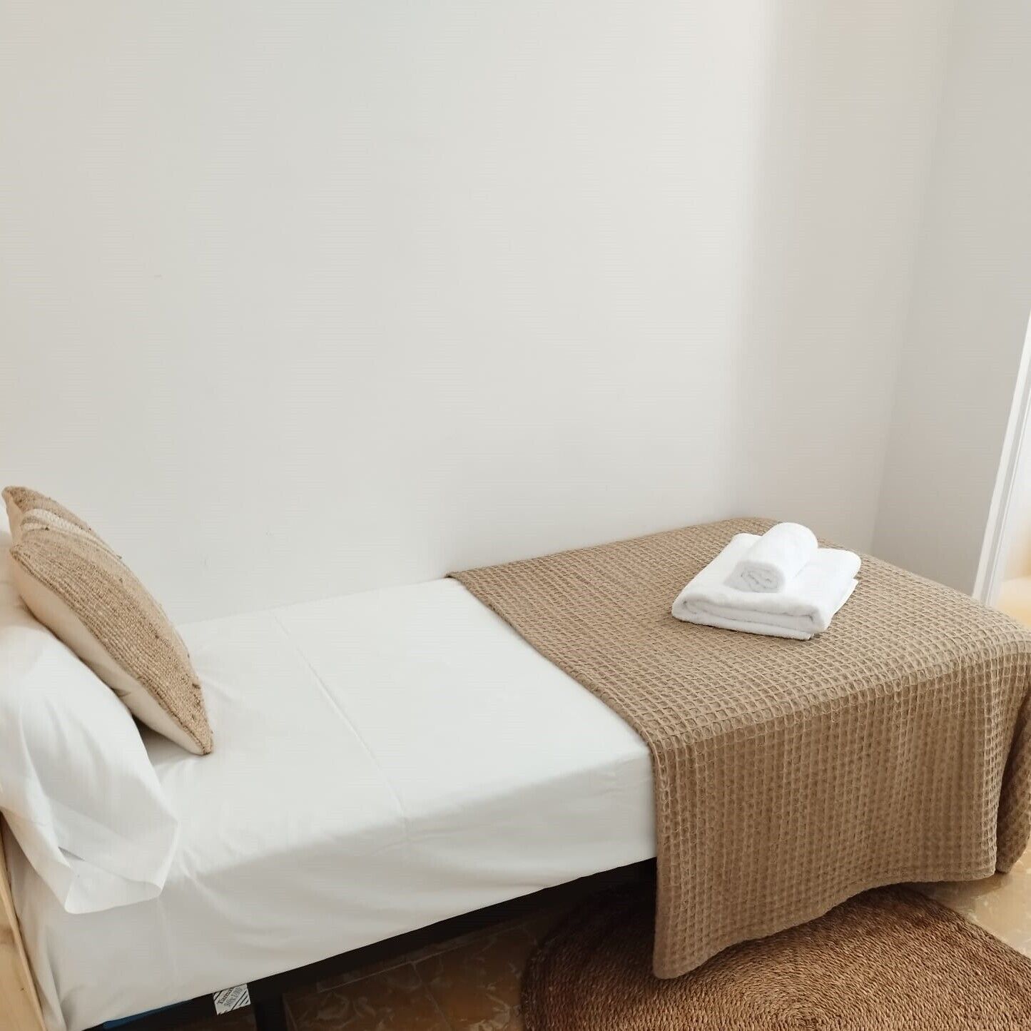 Una piccola camera con un letto singolo, un materasso bianco, un cuscino beige e una coperta marrone.