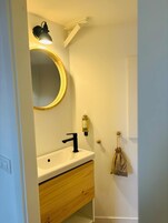 A bathroom with a white sink, black faucet, and a round mirror with a gold frame. There is a small bottle of soap on the wall and a towel hanging on a hook.