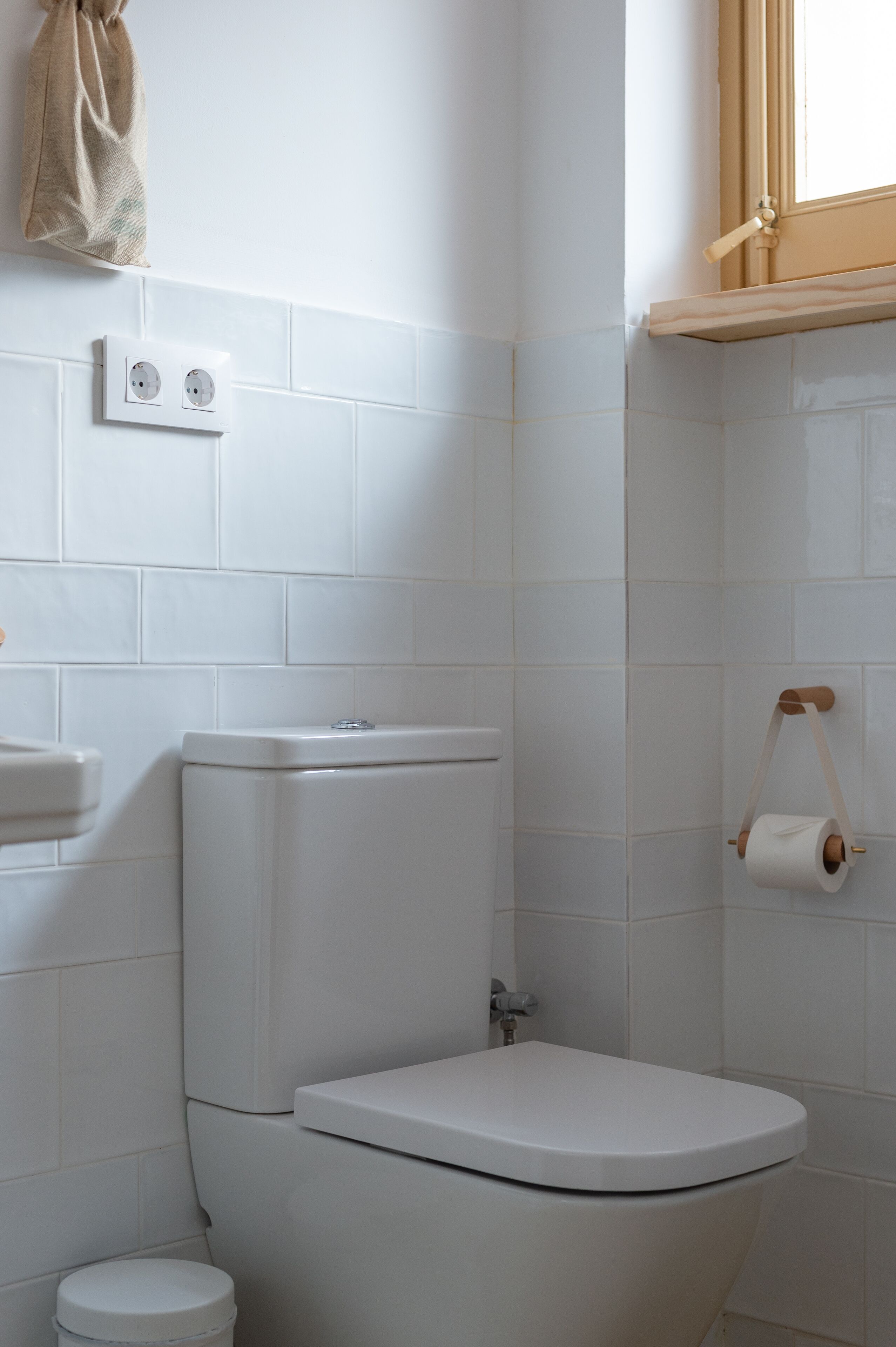 Un baño con azulejos blancos, inodoro, lavabo y ventana con persiana de madera.