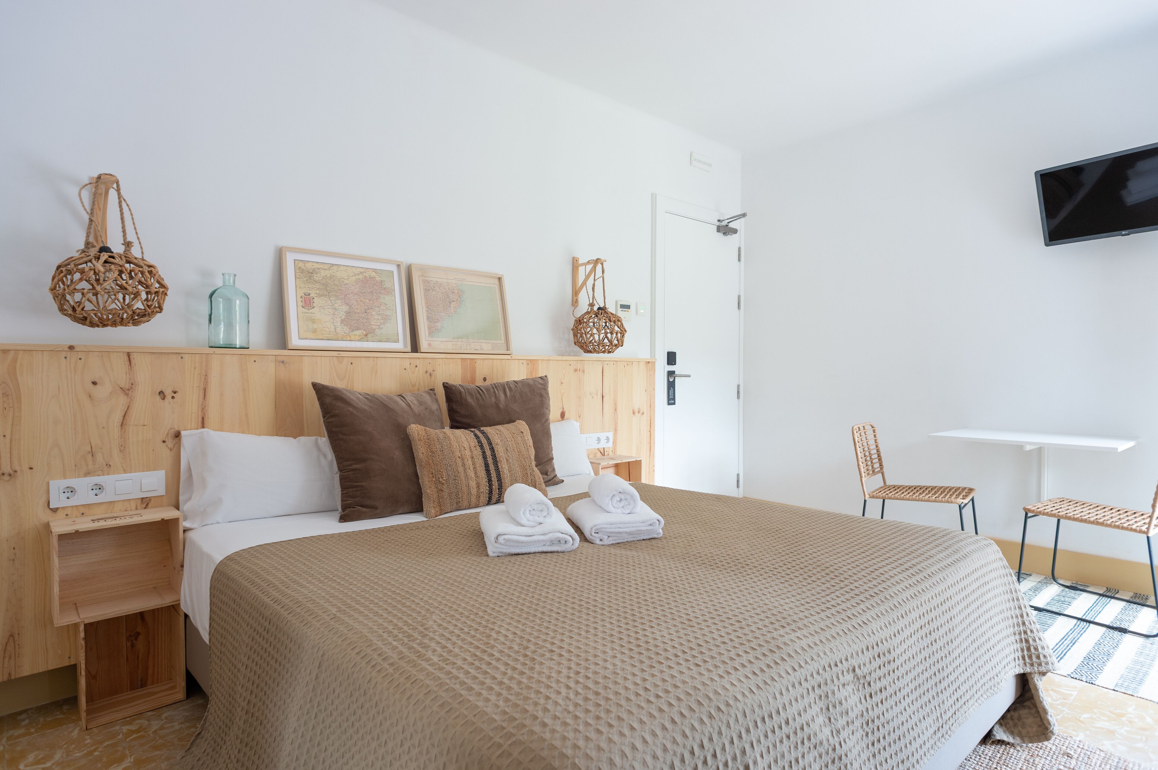 A neatly arranged hotel room with a wooden headboard, a bed with a beige blanket, two rolled towels, a small table, two wicker chairs, a wall-mounted TV, and a framed map on the wall.