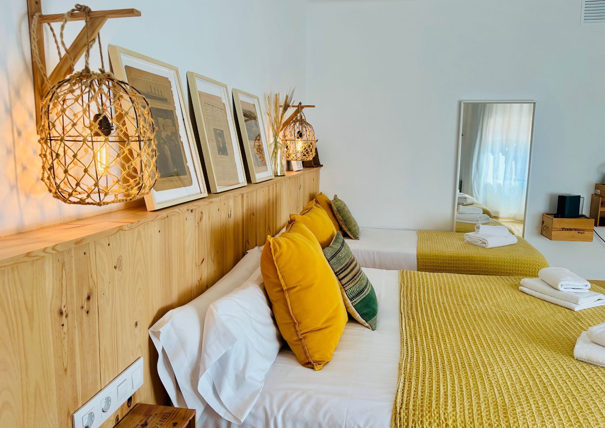 A hotel room with a wooden headboard, a bed with white and yellow bedding, a bedside table with a lamp, and a wall-mounted shelf with framed pictures and a decorative light.