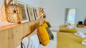A hotel room with a wooden headboard, a bed with white and yellow bedding, a bedside table with a lamp, and a wall-mounted shelf with framed pictures and a decorative light.
