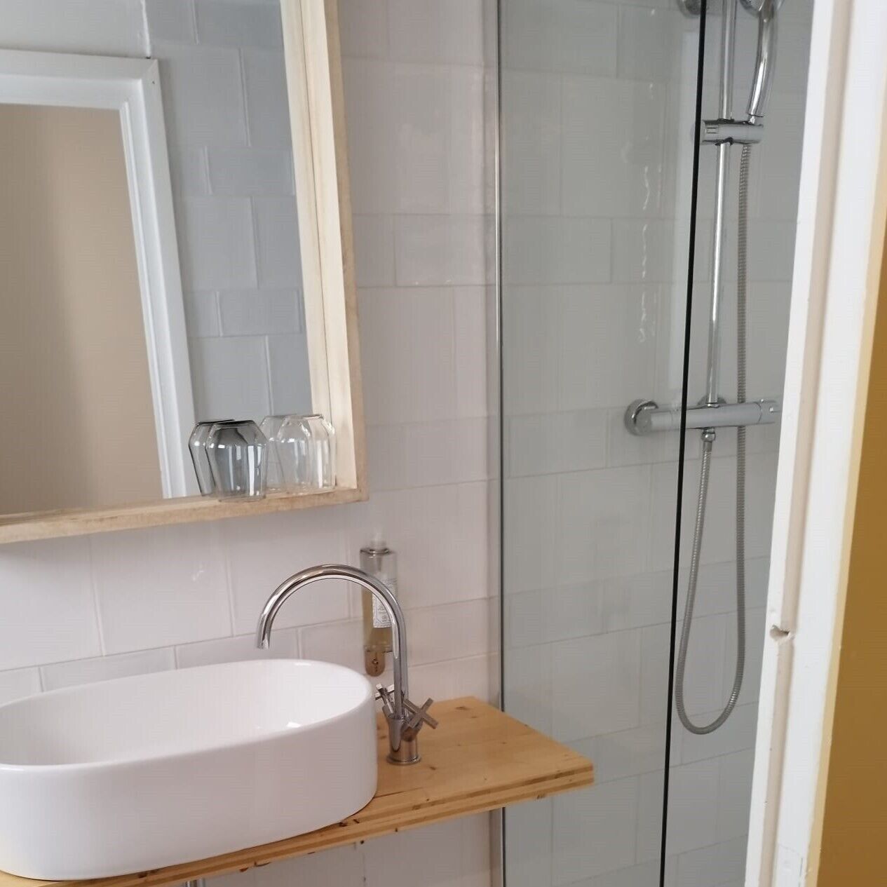 Un baño con un lavamanos blanco, un estante de madera, un espejo y una ducha con mampara de vidrio.