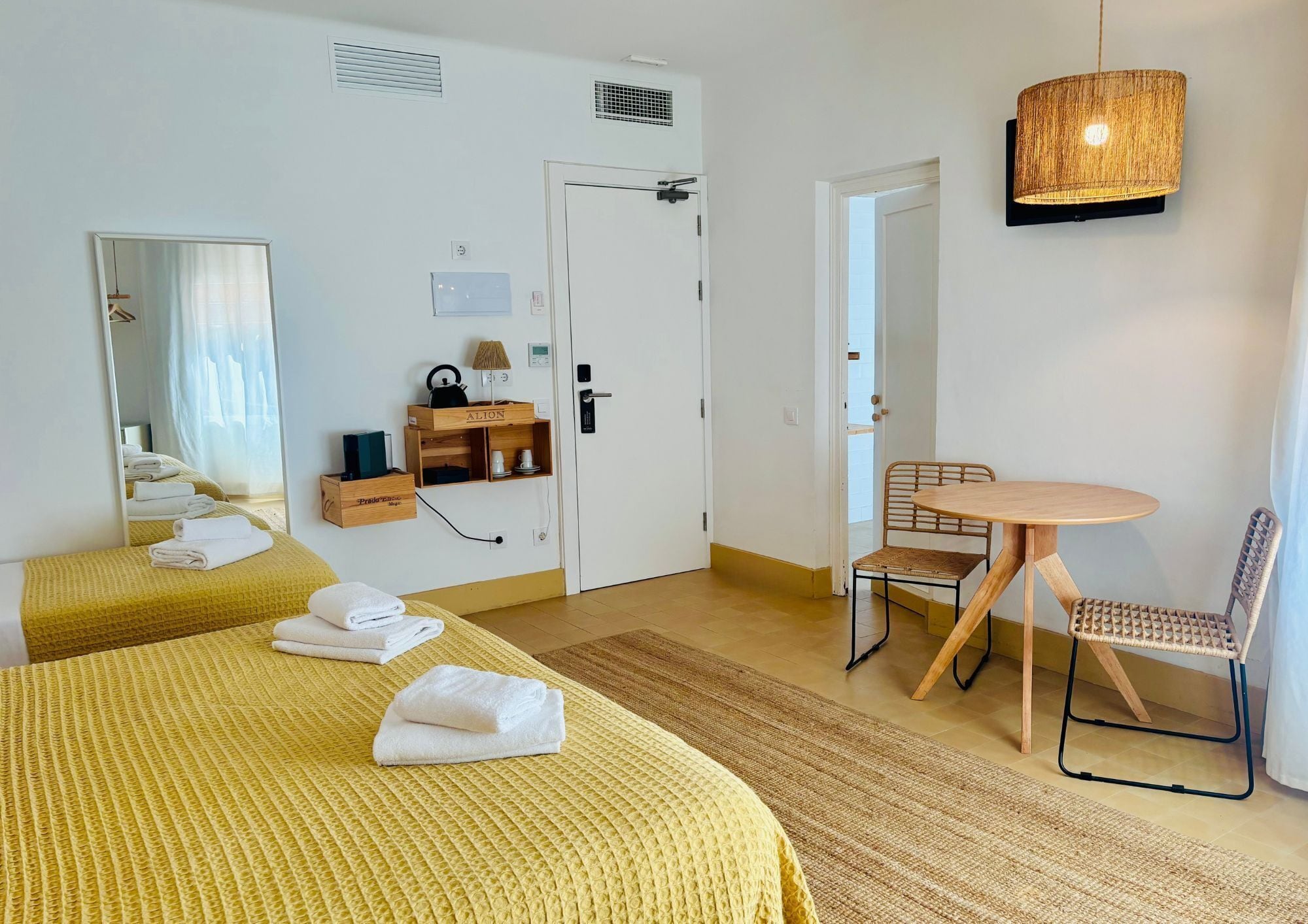 A hotel room with two beds, a small table with chairs, a mirror, a door, and a hanging light fixture.