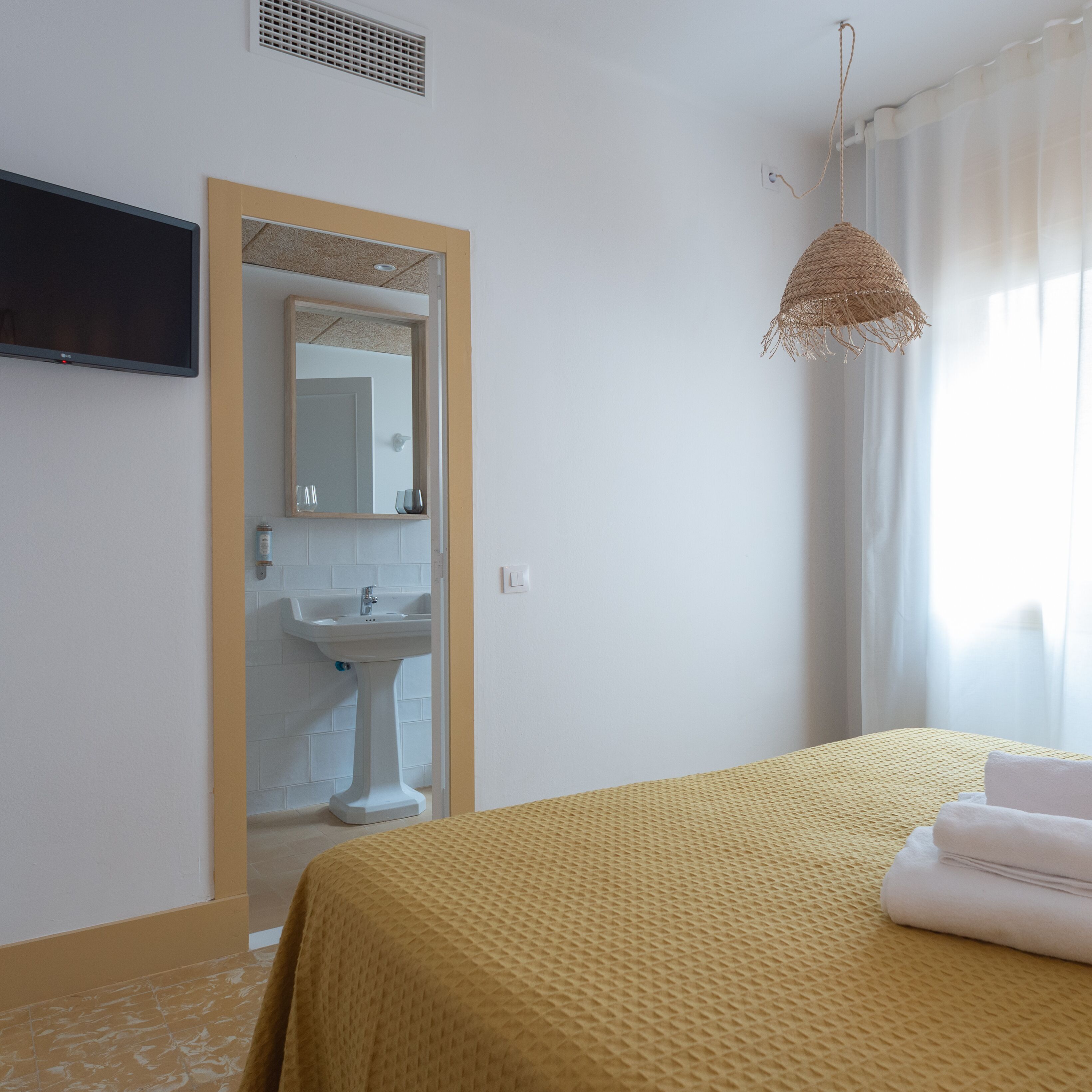 A bedroom with a bed, a bedside table, a mirror, a TV mounted on the wall, and a window with curtains.