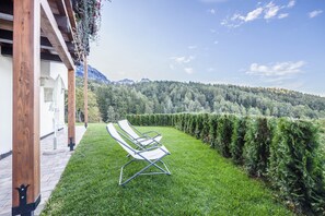A patio with two white lounge chairs, a wooden pergola, and a view of a forested mountain range.