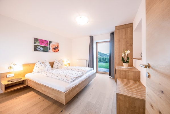 A modern bedroom with a wooden bed, bedside lamps, a view of greenery, and a wooden wall.