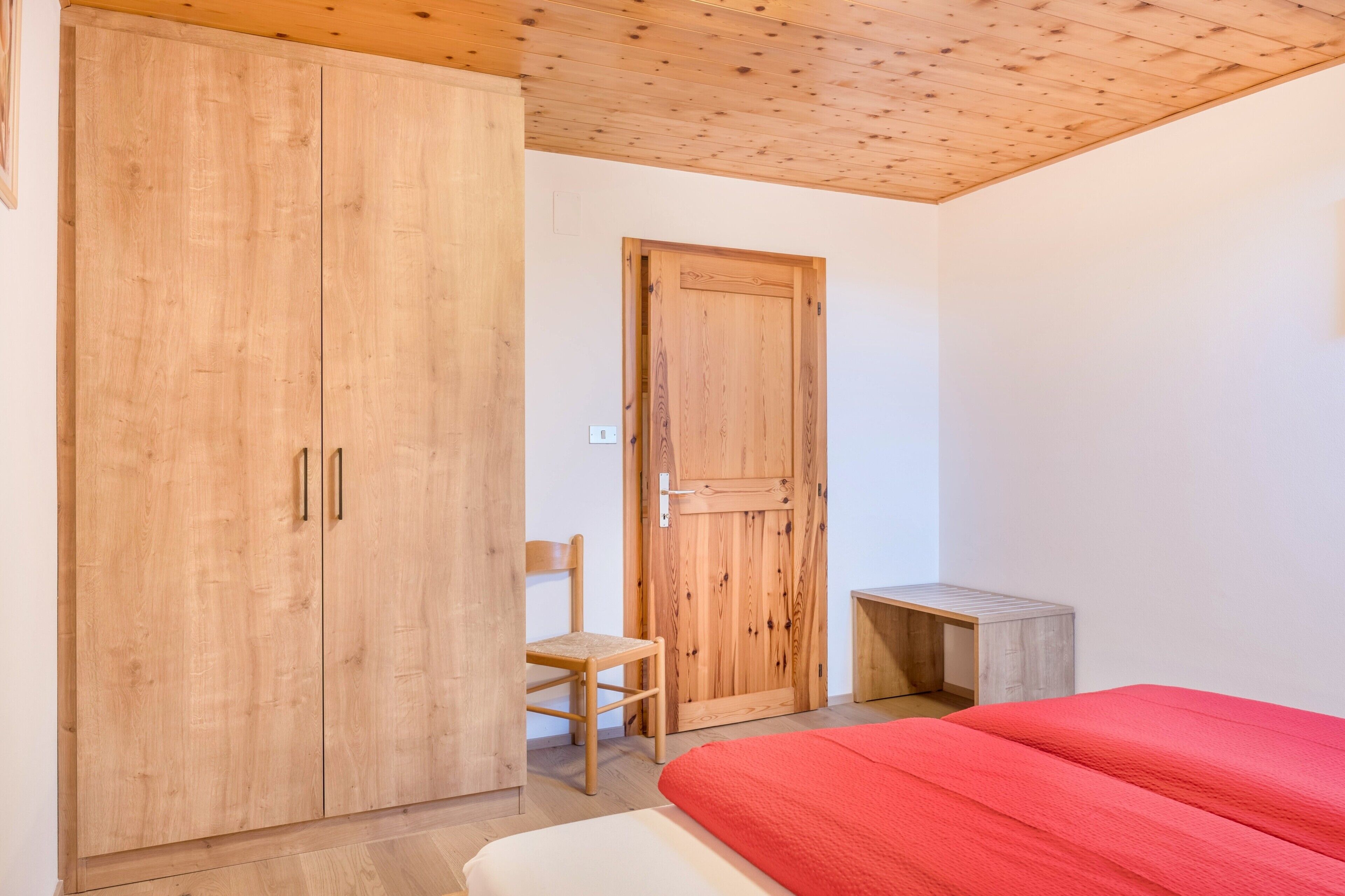 Un dormitorio con un armario de madera, una cama individual con una colcha roja, una silla de madera y una pequeña mesa de madera.