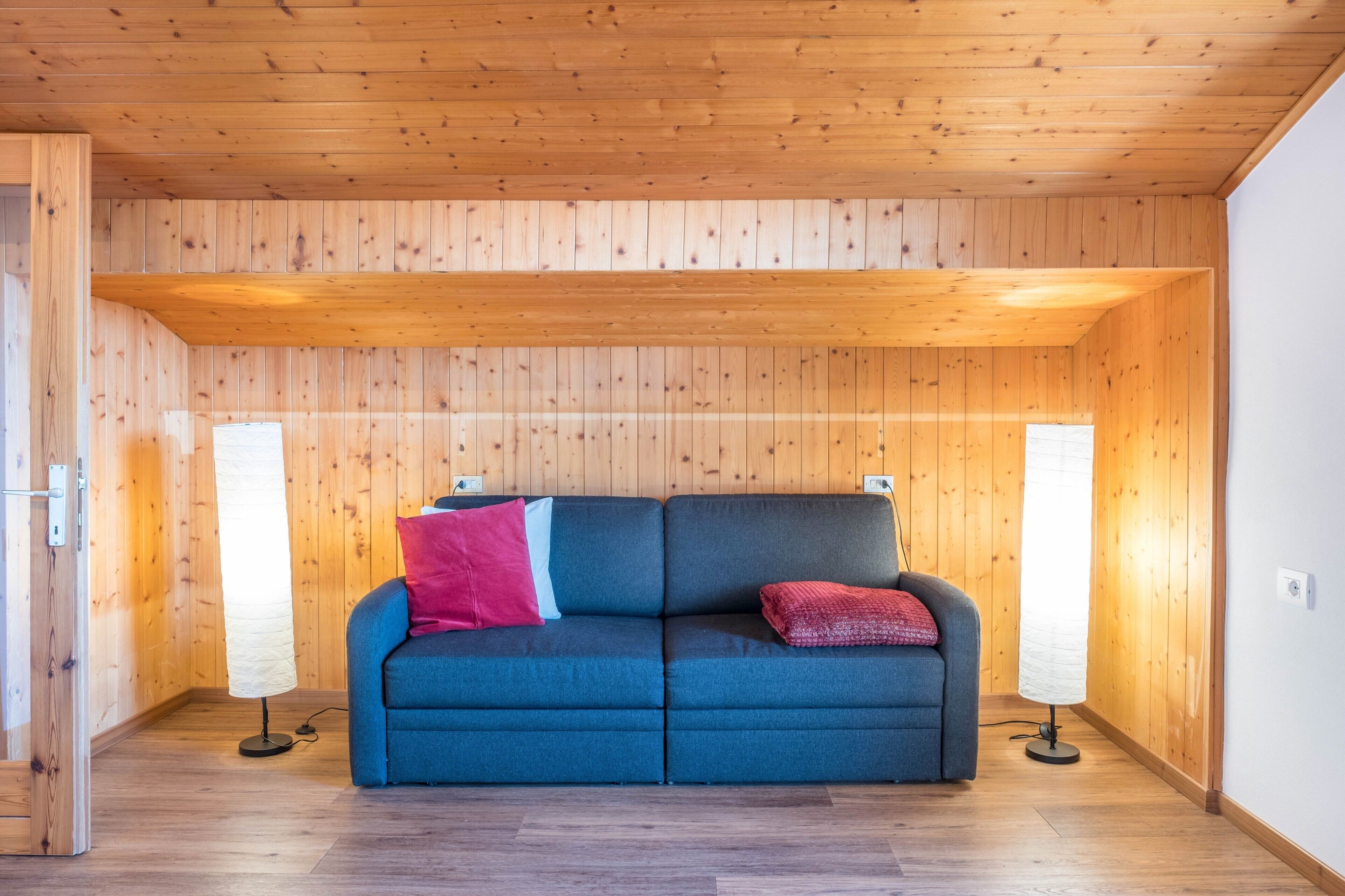 Een kamer met houten wanden en plafond, een blauwe bank met kussens, en twee vloerlampen.
