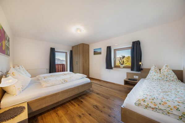 A bedroom with two beds, a wooden floor, a window with curtains, a bedside table, and a wall-mounted TV.