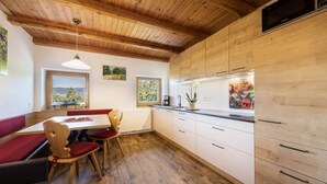 A kitchen with wooden cabinets, a dining table with chairs, and a view of the outdoors through windows. - Apartment “Ritten Gfliererhof” with Mountain View, Garden & Balcony (Völs am Schlern)