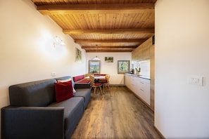 A compact living space with a wooden ceiling, a sofa with red cushions, a dining table with chairs, and a kitchen area with white cabinets.