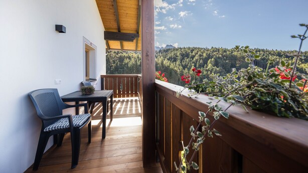 En balkon med et træbord og stole, udsigt til et skovklædt bjerglandskab, og gelænder af træ.