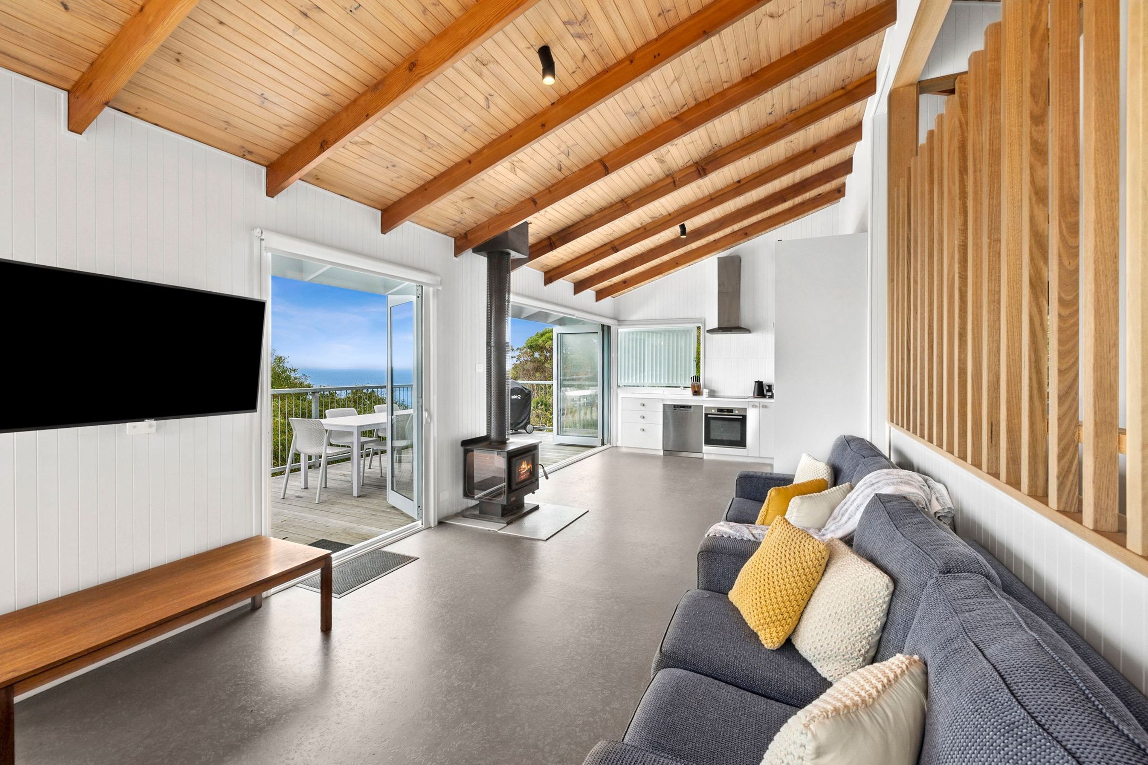 Ein modernes Wohnzimmer mit einem großen Fernseher, einem grauen Sofa mit Kissen und einer Holzbalkendecke.
