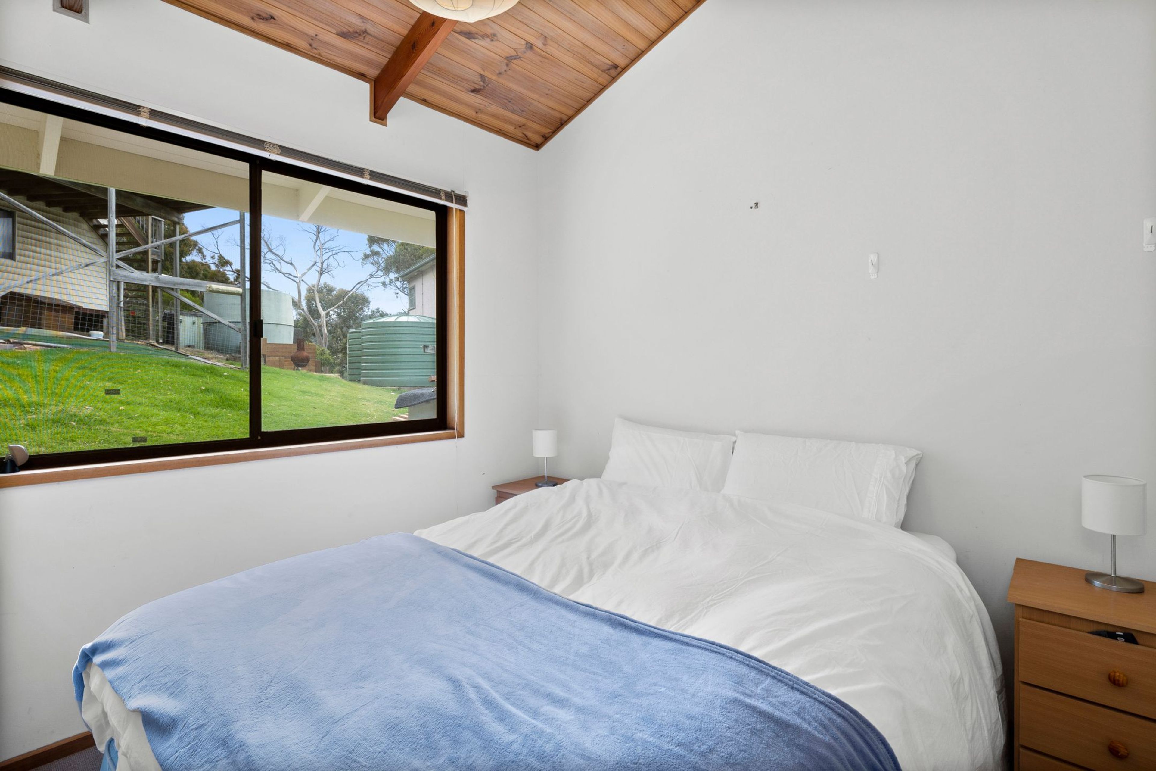 A bedroom with a bed, a nightstand, a lamp, and a window with a view of a yard and a building.