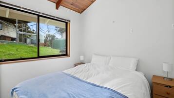 A bedroom with a bed, a nightstand, a lamp, and a window with a view of a yard and a building.