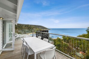 A balcony with a white table and chairs, overlooking a coastal view with a clear sky.