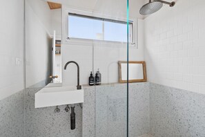A modern bathroom with a white sink, a shower with a rain showerhead, and a window above the sink.