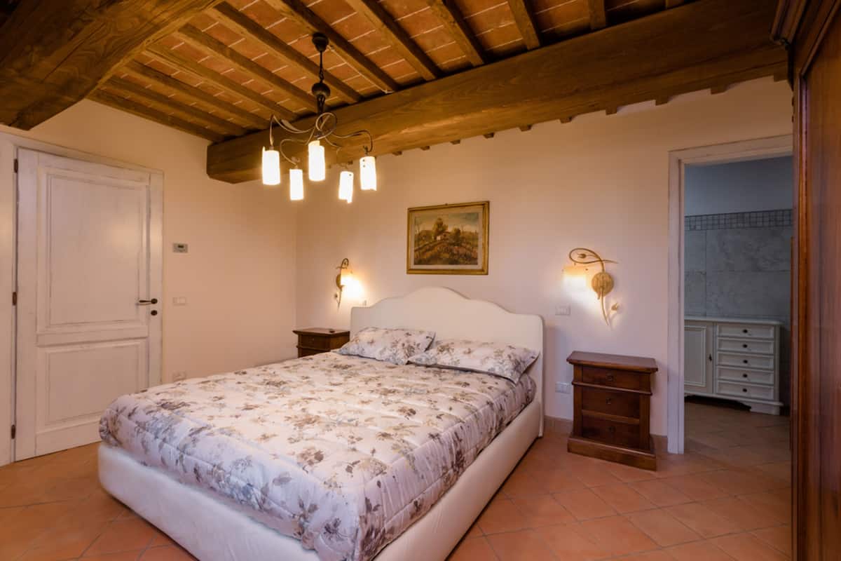 A bedroom with a wooden ceiling, a bed with floral bedding, two bedside tables with wall-mounted lamps, and a door.
