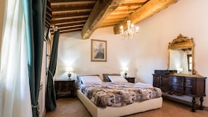 A bedroom with a large bed, wooden ceiling beams, a chandelier, a dresser, and a mirror. - Tenuta Tegolato (Barberino Tavarnelle)