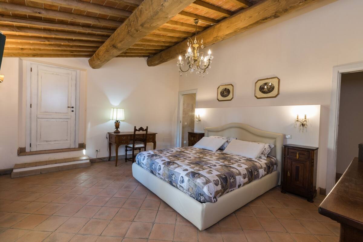 A bedroom with a bed, a desk, a chair, a chandelier, and a door.