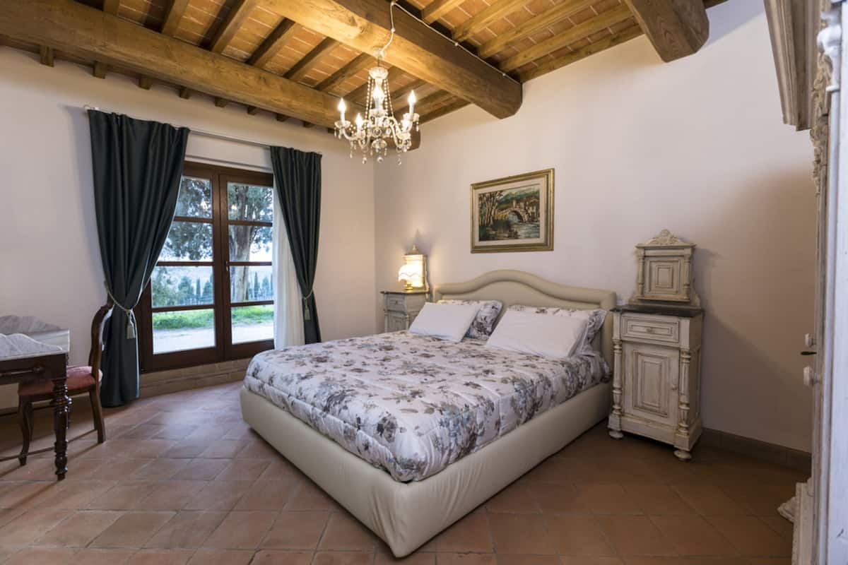 A bedroom with a large bed, a dresser, a chair, a window with curtains, and a chandelier.