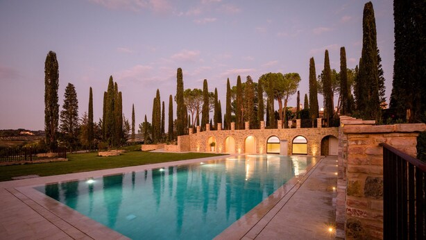 A rectangular swimming pool with a stone border, surrounded by a well-maintained lawn and tall trees. - Tenuta Tegolato (Barberino Tavarnelle)