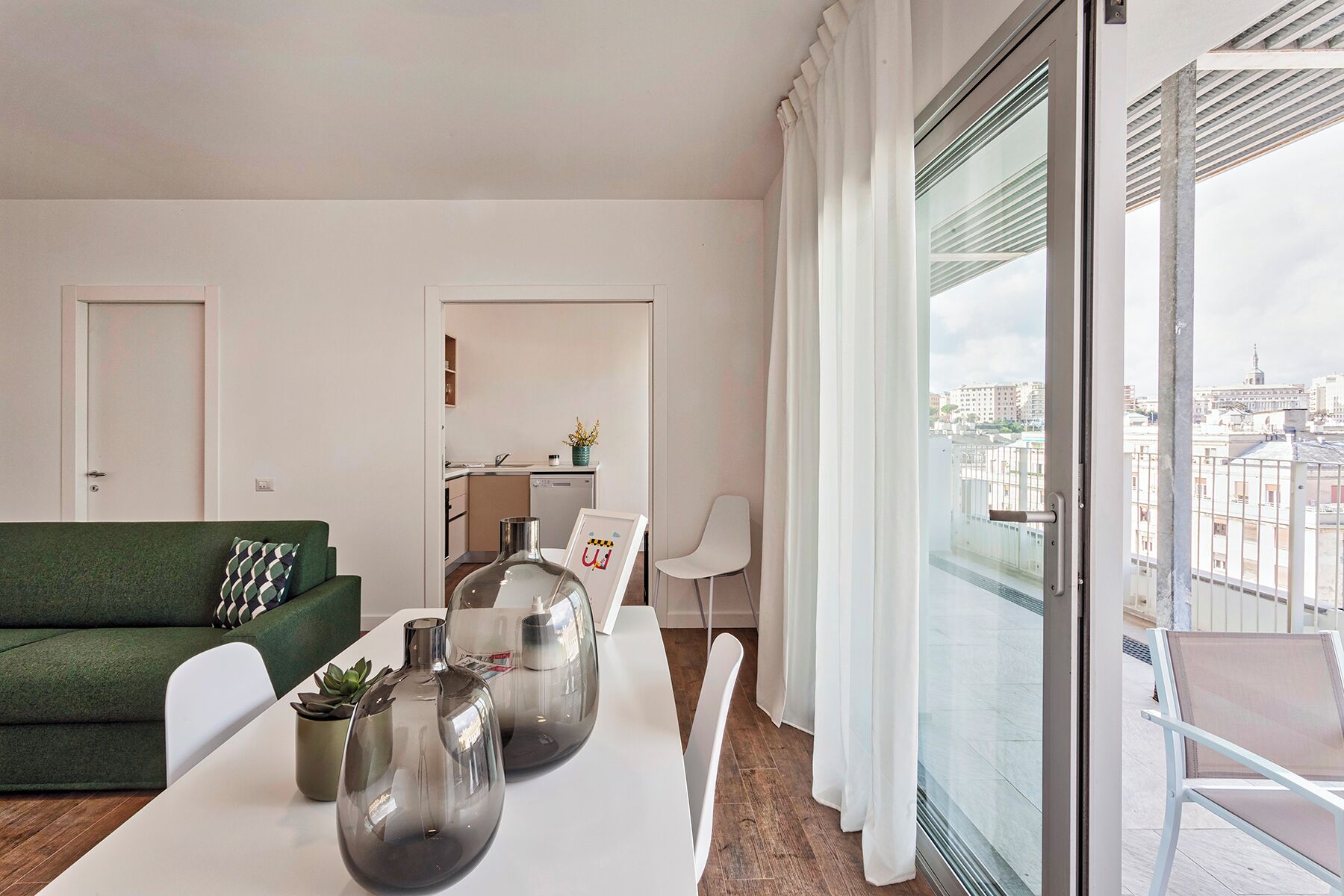 Una sala moderna con un sofá verde, sillas blancas y una mesa de comedor con jarrones decorativos. Se ve una cocina al fondo, y un balcón con vista a la ciudad a la derecha.
