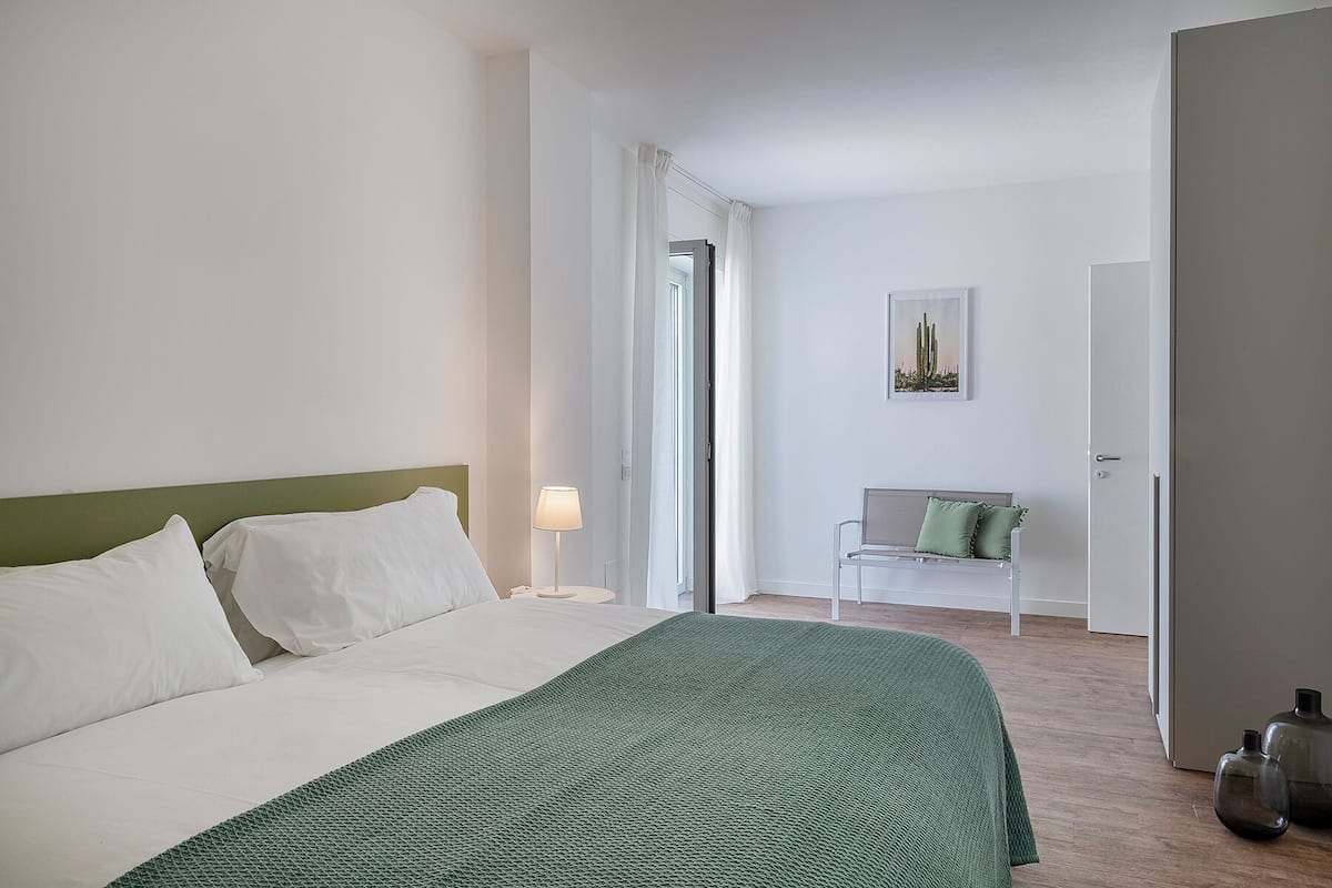 A hotel room with a bed, a green throw, a nightstand, a lamp, a chair, and a door leading to another room.