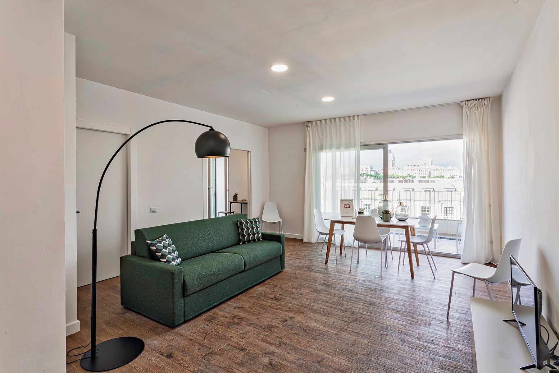 Un salon moderne avec un canapé vert, une table à manger avec des chaises, un lampadaire et une grande fenêtre avec des rideaux.