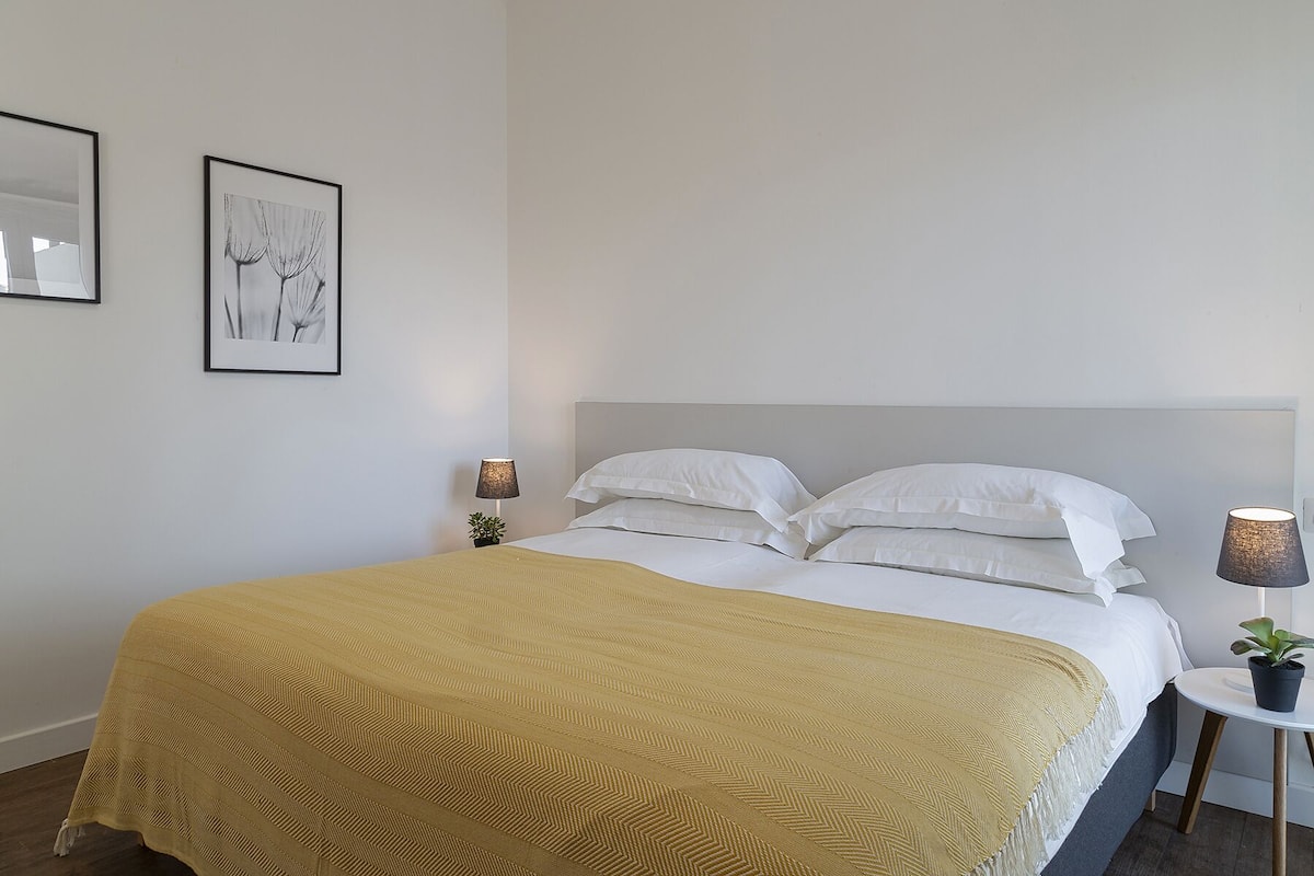 A neatly made bed with white linens and a mustard-colored blanket, a bedside lamp, and two framed pictures on the wall.