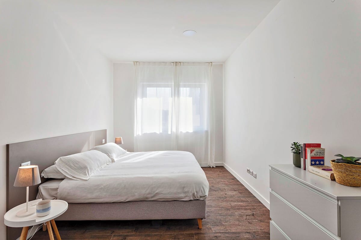 A bedroom with a bed, bedside table, lamp, and a dresser with a basket and books.
